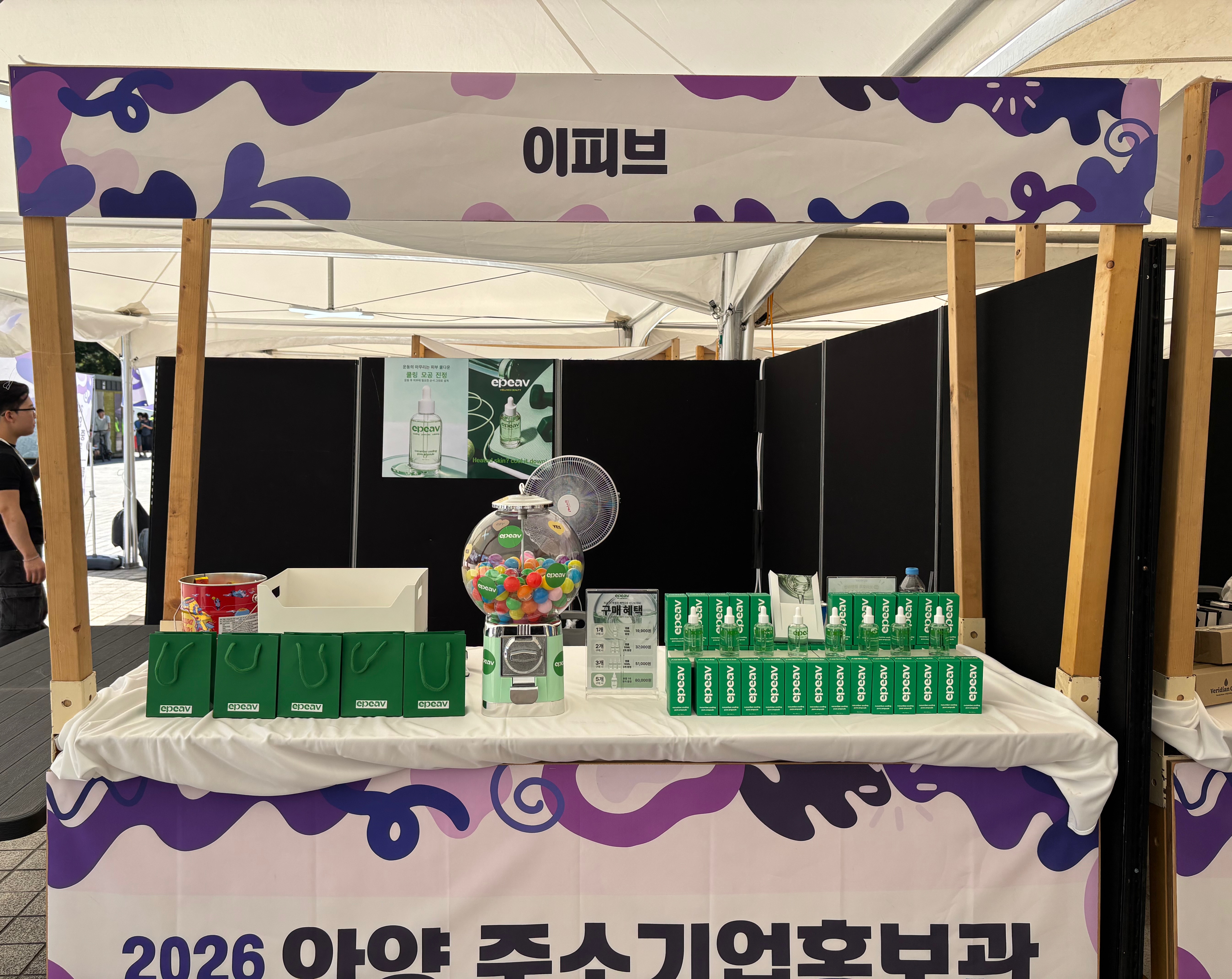 보라색 그래픽 배너 아래 '이피브' 간판이 걸린 부스에 초록 쇼핑백, 뽑기 기계, 앰플 진열대가 놓여 있다.