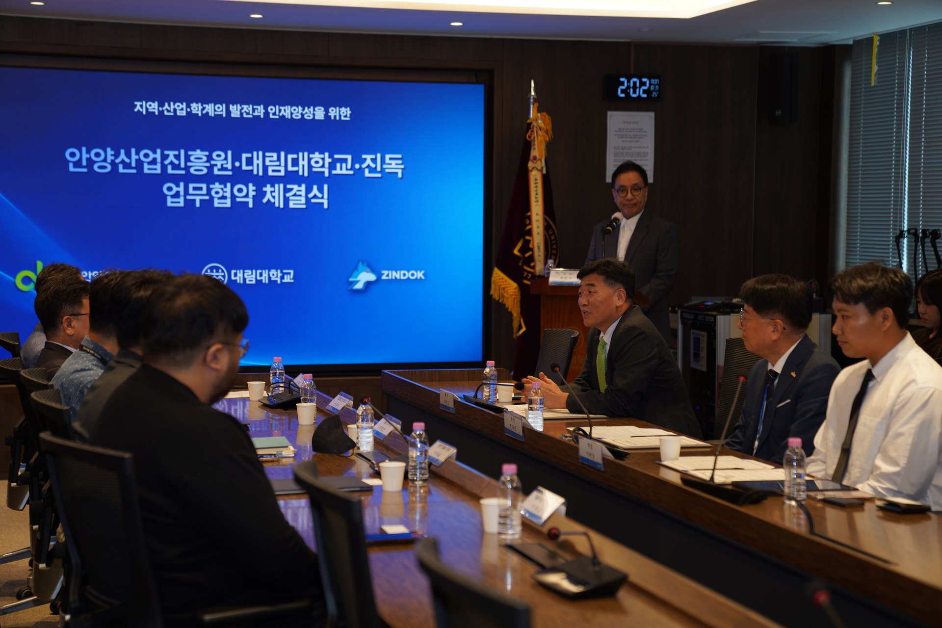 회의 테이블에 앉은 초록 넥타이 남성이 마이크 앞에서 말하고 맞은편 참석자들이 듣고 있다