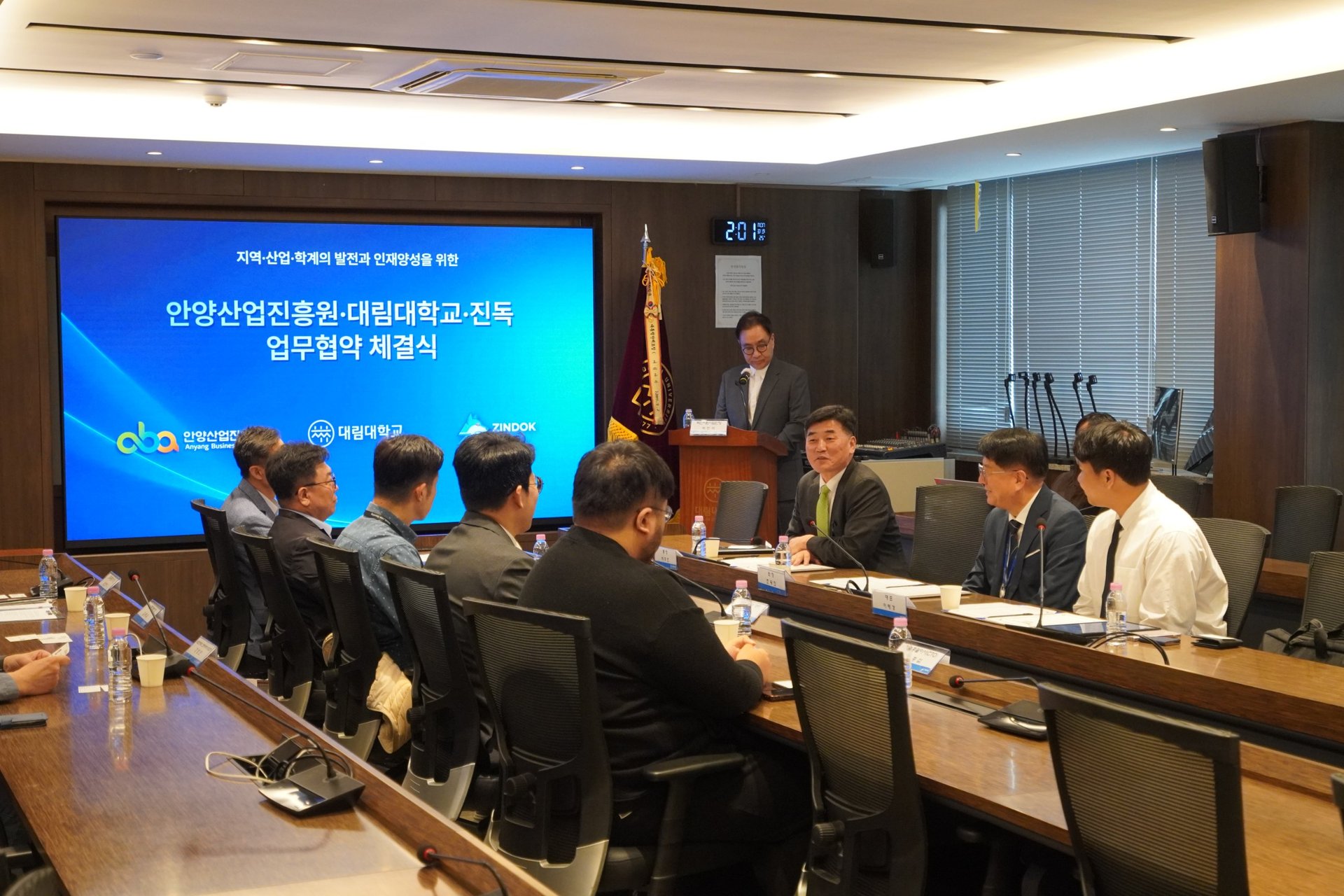안경 쓴 정장 차림 남성이 회의 테이블에서 말하고 양옆의 두 남성이 서류를 앞에 두고 앉아 있다
